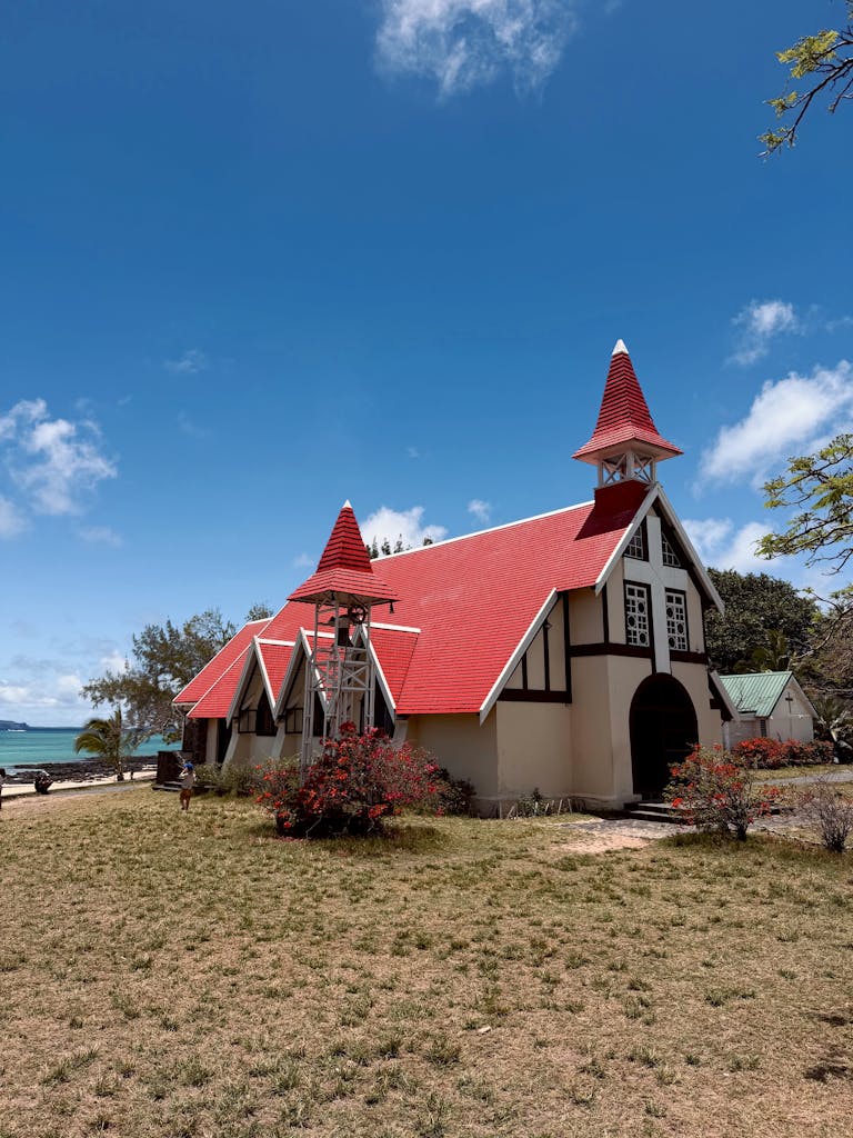 Picturesque church with red roof by the sea under bright blue sky and lush greenery.